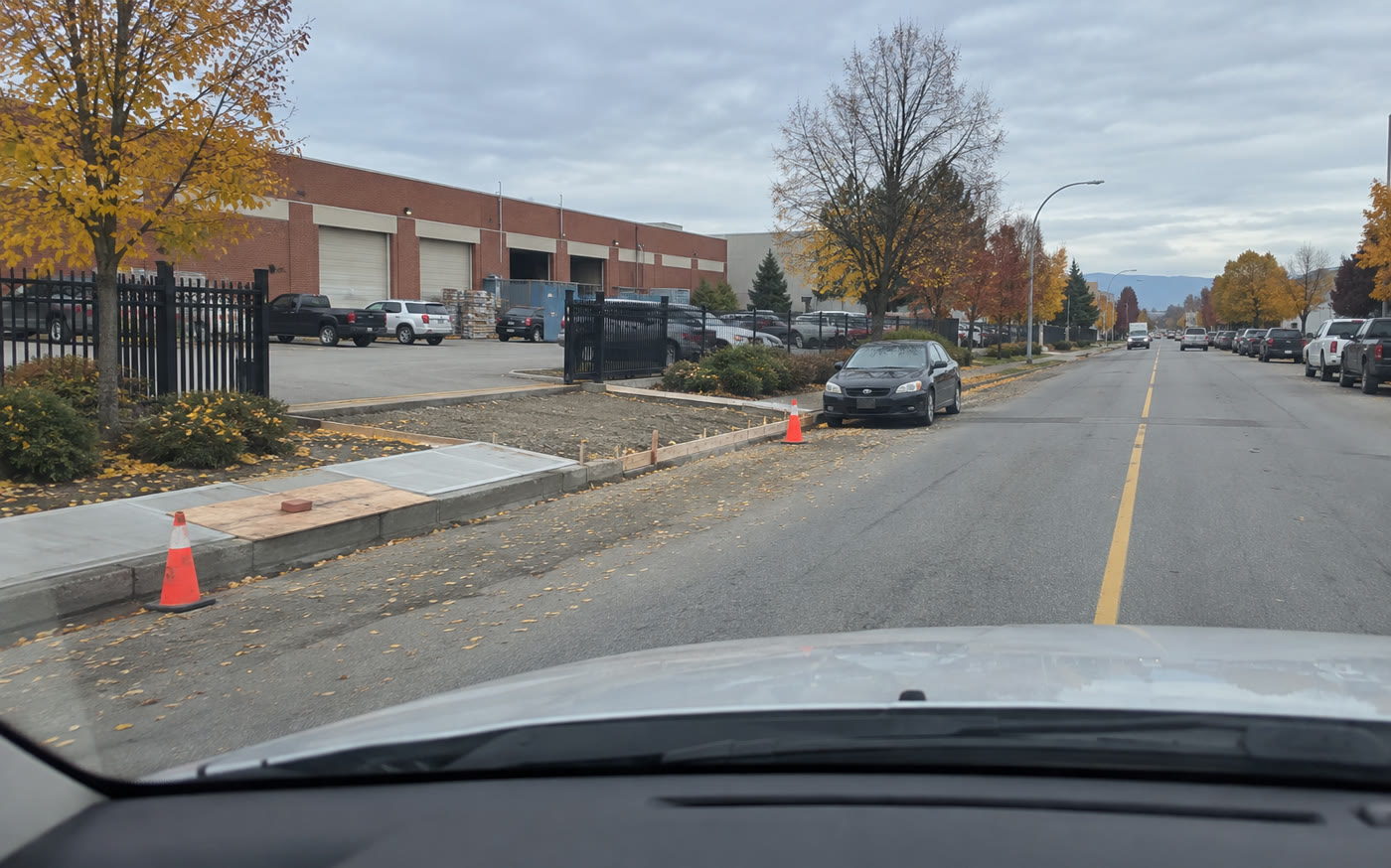 From the lane in autumn on a brick-fronted industrial street: two cones at the curb, a new sidewalk panel with plywood held down by a brick, and a driveway apron formed on gravel base, not yet poured, a car parked at the curb beside it